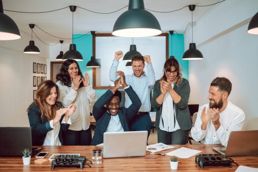 Group of cheerful diverse colleagues clapping and celebrating success while gathering at table with laptop and documents in modern office